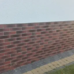 Ceglana okładzina elewacyjna w odcieniach czerwieni i brązu, kontrastująca z jasną, gładką ścianą powyżej i kostką brukową u podstawy. Widoczny fragment trawnika.