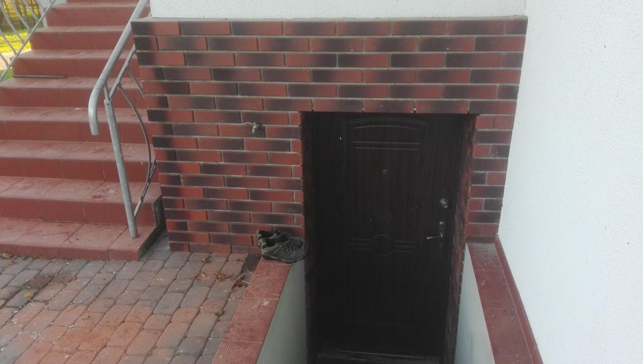 Ciemne drzwi wejściowe w obramowaniu z cegły klinkierowej, z parą butów leżących obok, widoczne schody wejściowe i fragment białej ściany.