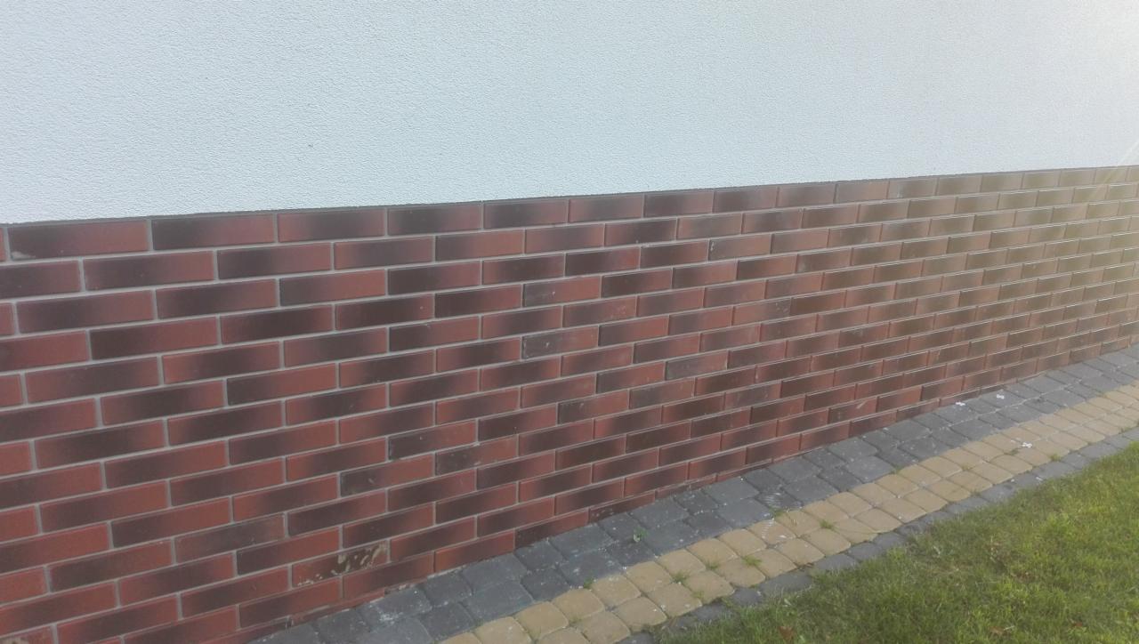 Ceglana okładzina elewacyjna w odcieniach czerwieni i brązu, kontrastująca z jasną, gładką ścianą powyżej i kostką brukową u podstawy. Widoczny fragment trawnika.