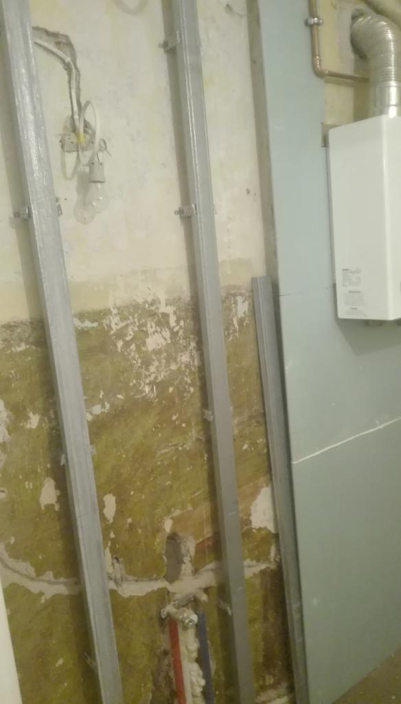 Montaż stalowych profili na nierównej ścianie z widocznymi instalacjami elektrycznymi i rurami, przygotowanie pod obłożenie płytami gipsowo-kartonowymi, w tle biały piec gazowy.