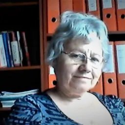 Portret kobiety w okularach na tle regału z książkami i segregatorami, uśmiechająca się do kamery.