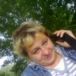 Uśmiechnięta kobieta z blond pasemkami rozmawia przez telefon na tle zielonych drzew.