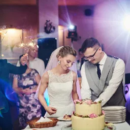 Para młoda kroi tort weselny, goście w tle trzymają zimne ognie, widoczny reflektor i tańcząca para.