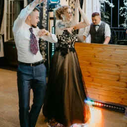 Para młoda w tańcu podczas wesela, panna młoda w sukni z czarną koronką, pan młody w eleganckim garniturze i wzorzystym krawacie, DJ w tle za drewnianym barem z oświetleniem LED.