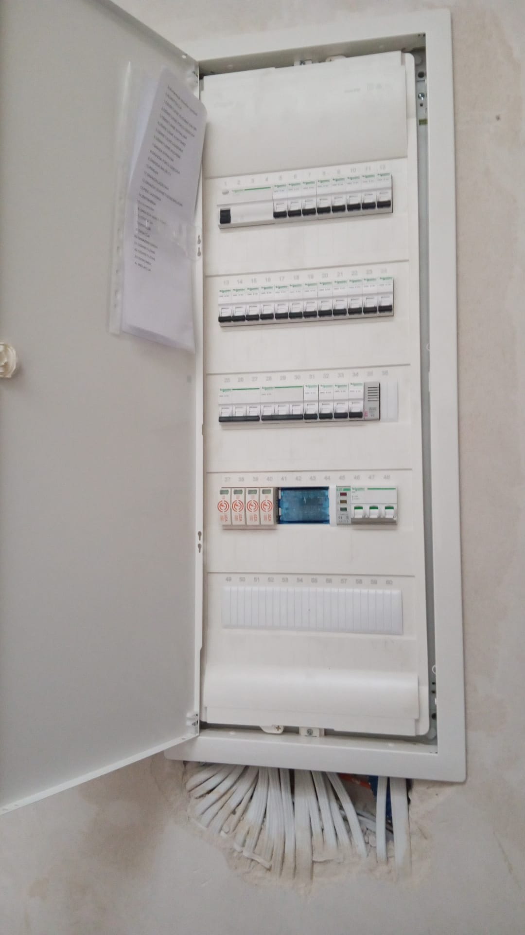Otwarta skrzynka rozdzielcza z widocznymi bezpiecznikami i przewodami elektrycznymi w ścianie, dokumentacja w foliowej koszulce przyczepiona do drzwiczek.