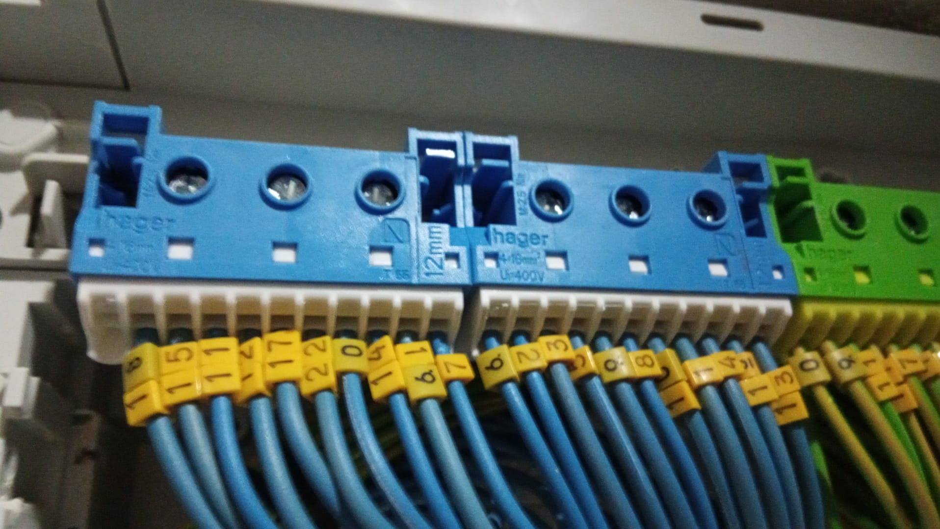 Szczegółowe ujęcie tablicy rozdzielczej z niebieskimi i zielonymi listwami zaciskowymi firmy Hager, do których podłączone są ponumerowane, niebieskie i zielono-żółte przewody.