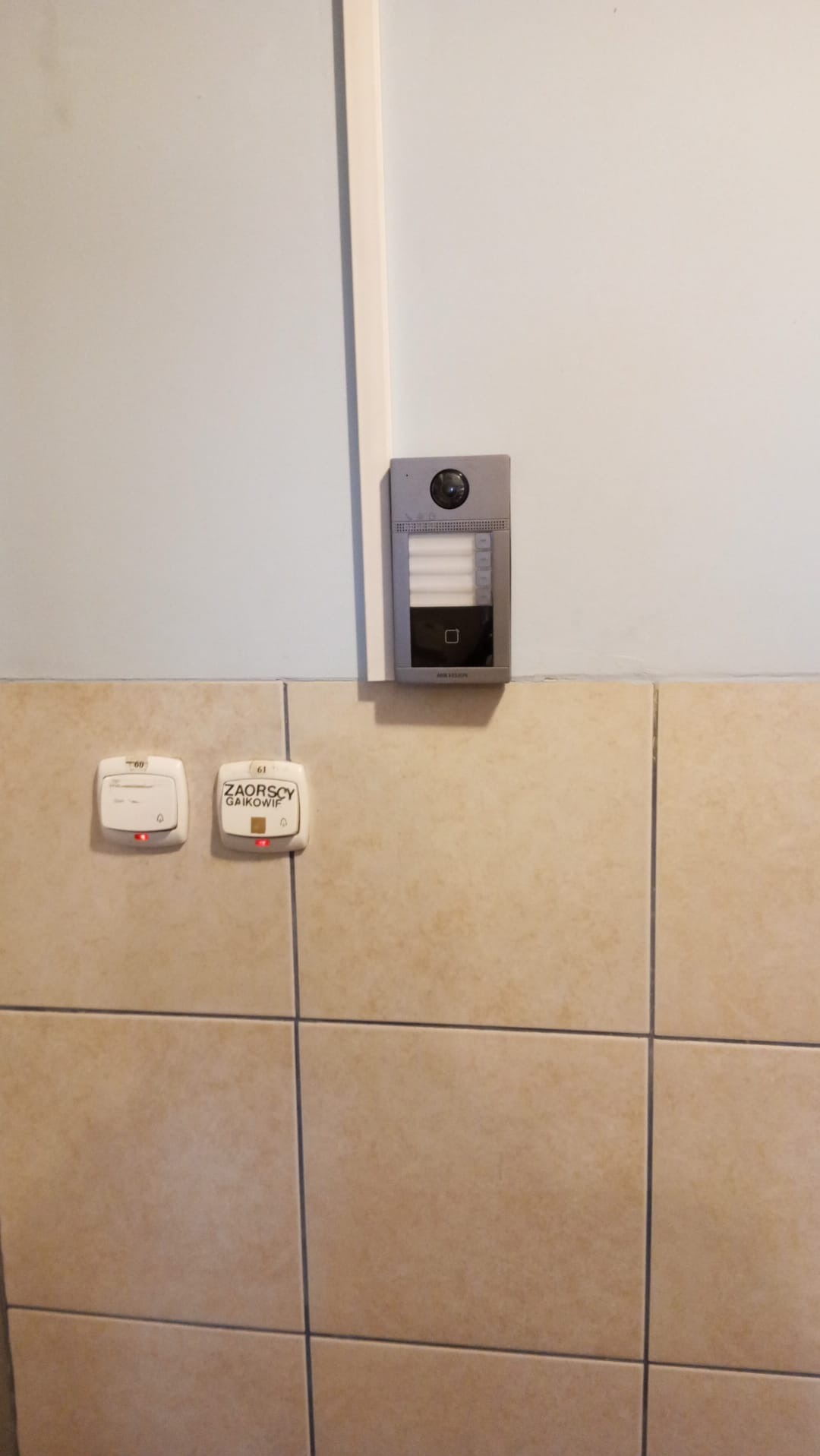 Zamontowany wideodomofon Hikvision na ścianie z beżowych płytek, obok dwa przyciski dzwonkowe z czerwonymi diodami, widoczne okablowanie.