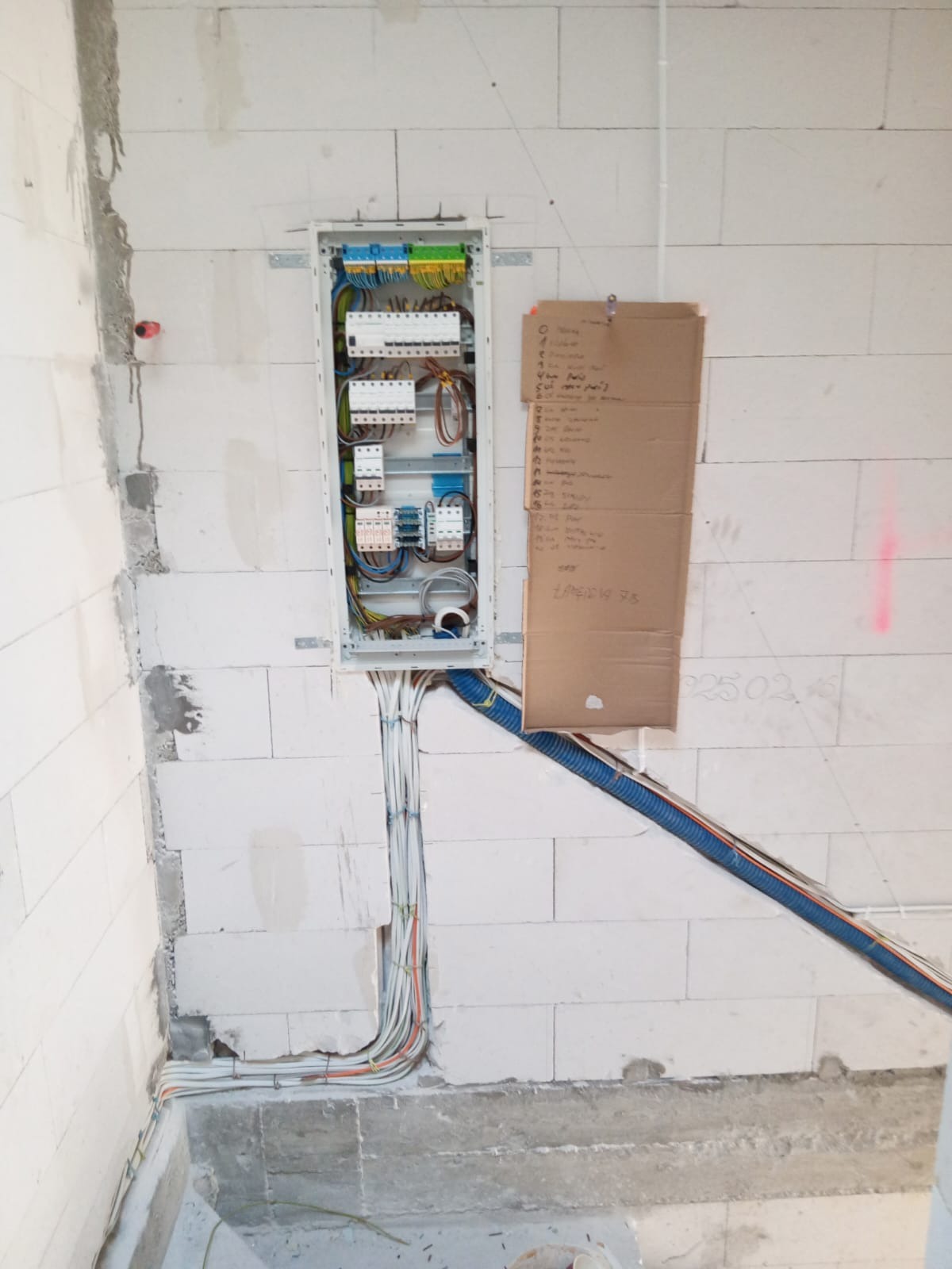 Rozdzielnia elektryczna z poprowadzonymi przewodami w ścianie z betonu komórkowego, widoczna niebieska rura karbowana i kartka z notatkami.