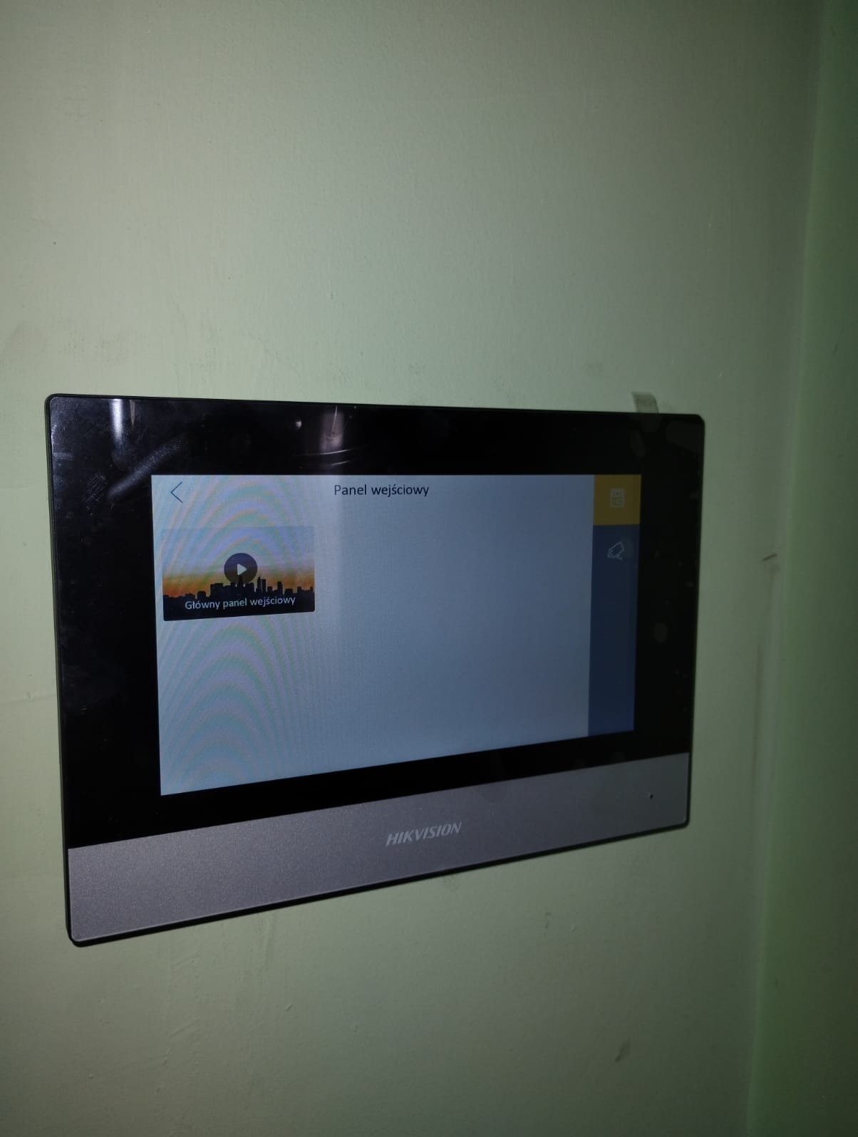 Zamontowany panel wideodomofonu Hikvision z widocznym menu 'Panel wejściowy' na ekranie dotykowym, zamocowany na zielonej ścianie. Widoczny fragment taśmy klejącej nad urządzeniem.