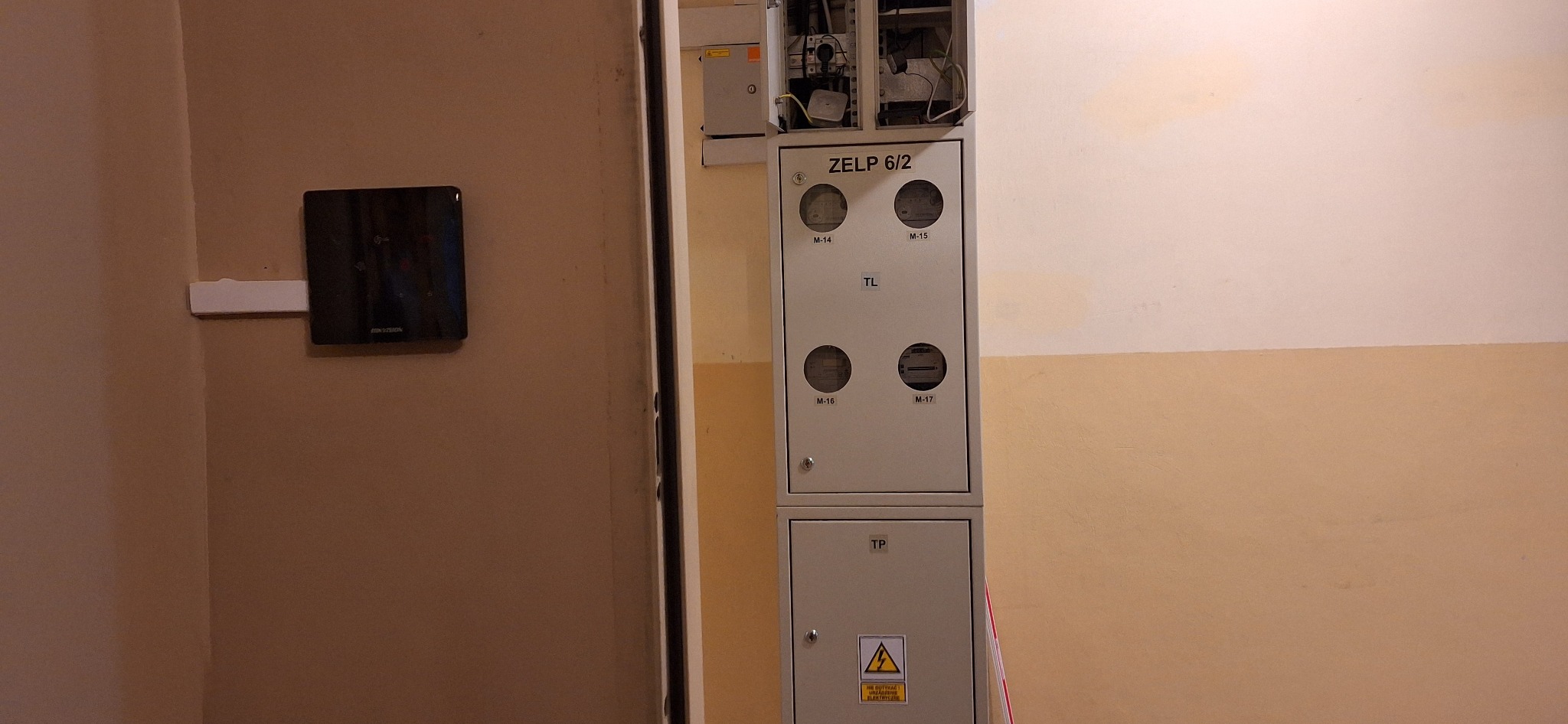 Szara skrzynka elektryczna ZELP 6/2 z czterema licznikami, poniżej skrzynka TP z symbolem ostrzegawczym o napięciu, na ścianie obok czarny panel kontrolny dostępu Hikvision.