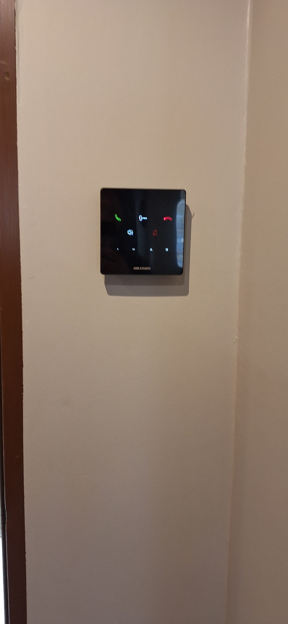 Czarny panel dotykowy wideodomofonu Hikvision zamontowany na beżowej ścianie przy brązowej futrynie drzwi, z podświetlonymi ikonami połączenia, głośnika i klucza.