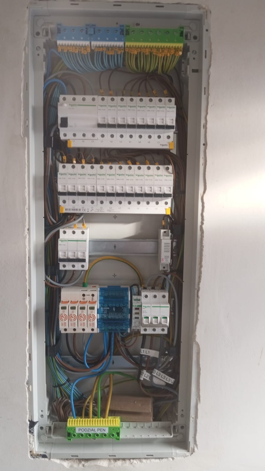 Rozdzielnica elektryczna z zainstalowanymi bezpiecznikami, zabezpieczeniami przepięciowymi i listwą PEN, widoczne okablowanie i oznaczenia faz.