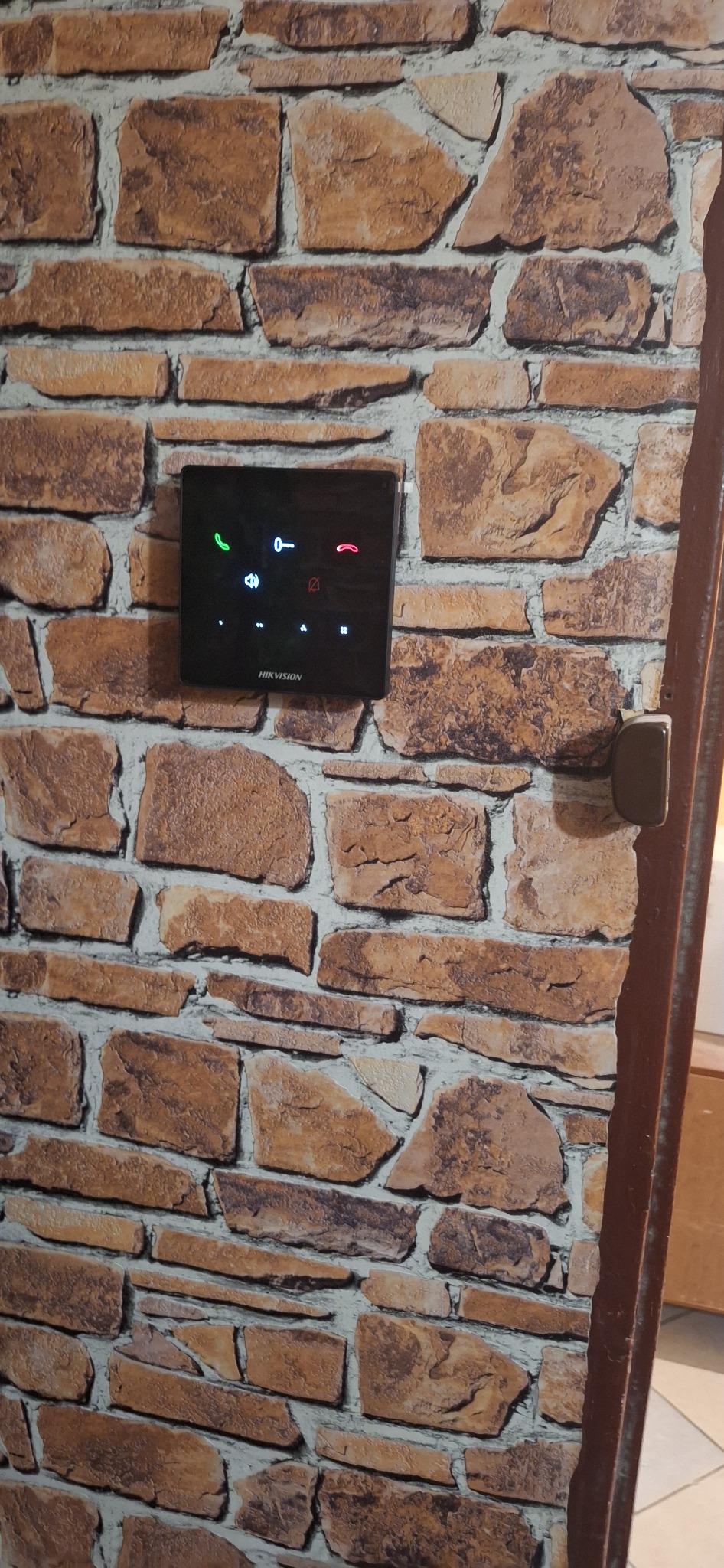 Czarny panel dotykowy domofonu Hikvision zamontowany na ścianie z tapetą imitującą kamień, widoczny fragment futryny po prawej stronie.