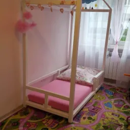 Białe łóżko domek dla dziecka z różowym materacem i poduszką w pokoju z kolorowym dywanem w motywy miejskie, ozdobione girlandą serc i chmurką.