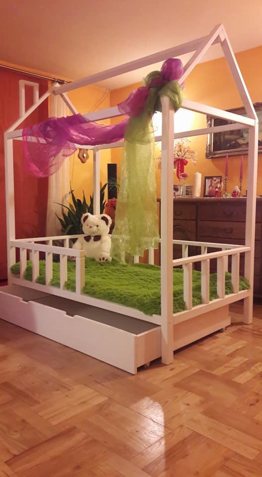Białe łóżko w kształcie domku z wysuwaną szufladą, zielonym futrzanym materacem i dekoracyjnymi tiulami w kolorze fuksji i limonki, pluszowy miś w środku.