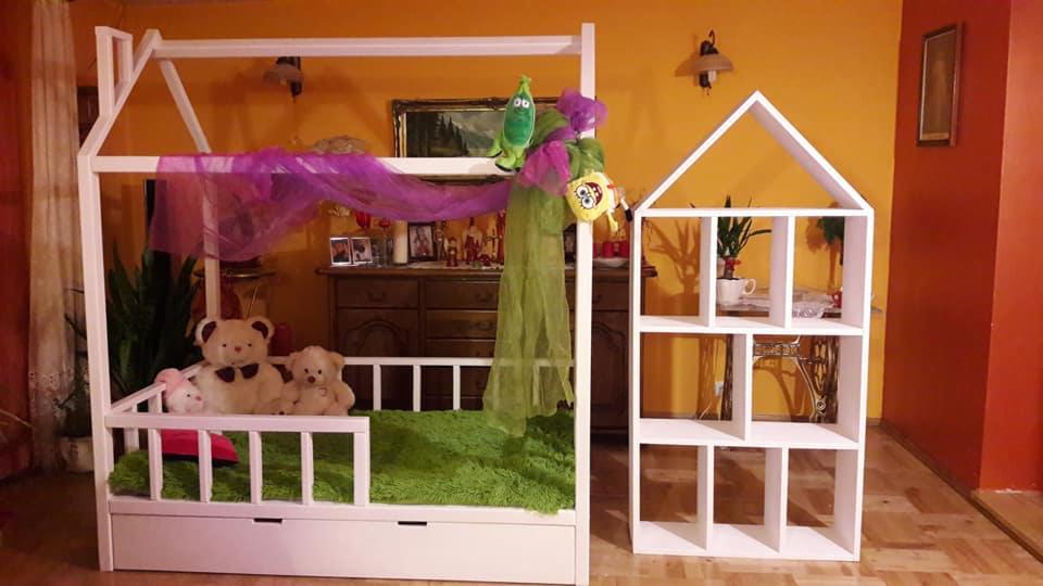 Białe łóżko domek dla dziecka z pluszakami i baldachimem, obok biały regał domek na zabawki, na tle pomarańczowej ściany.