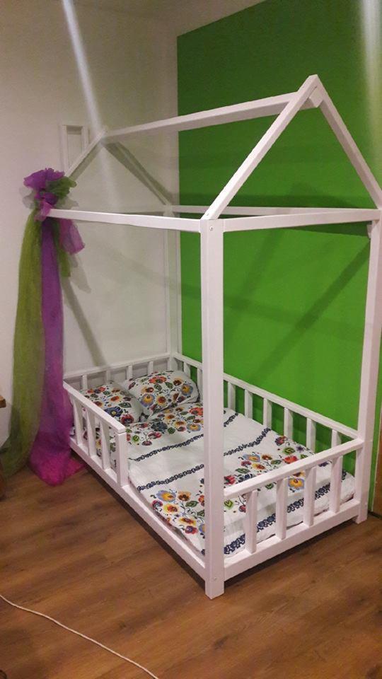 Białe łóżko dziecięce w kształcie domku z barierkami, zdobione fioletowo-zielonym tiulem, z pościelą w kwiaty, na tle zielonej ściany i drewnianej podłogi.