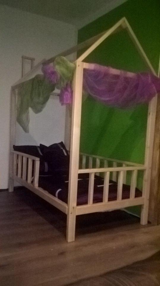 Drewniane łóżko dziecięce w kształcie domku z moskitierą w odcieniach zieleni i fioletu, ustawione na podłodze z paneli w pokoju z zieloną ścianą.