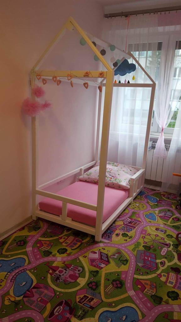 Białe łóżko domek dla dziecka z różowym materacem i poduszką w pokoju z kolorowym dywanem w motywy miejskie, ozdobione girlandą serc i chmurką.