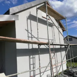 Dom w Częstochowie w trakcie prac elewacyjnych, widoczne rusztowania, świeżo nałożony jasnoszary tynk oraz fragmenty czarnej folii ochronnej na trawniku.