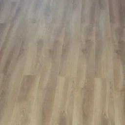 Ułożona pod kątem podłoga z paneli imitujących drewno o jasnym odcieniu, widoczna przy krawędzi ściany z białym cokołem.