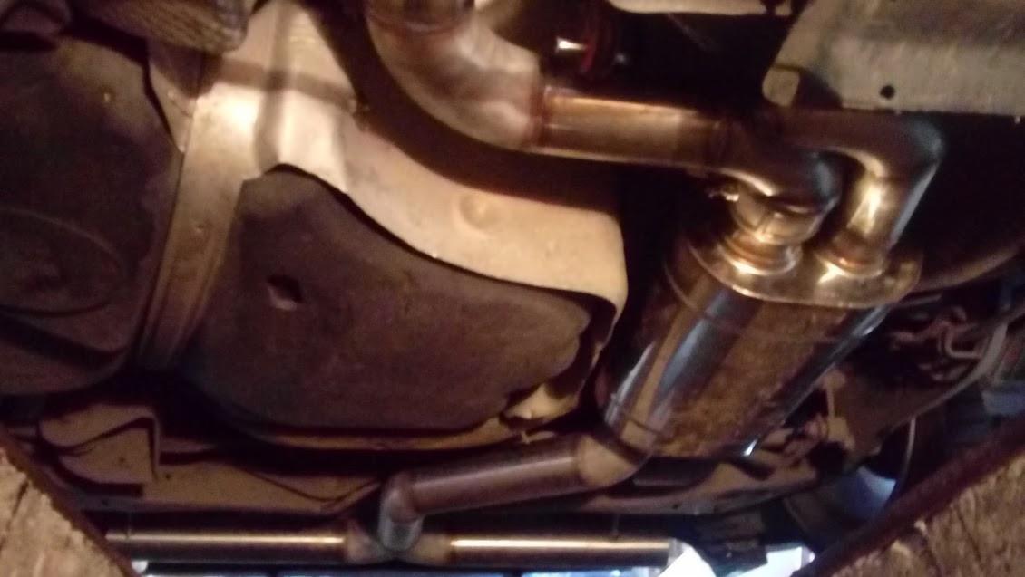 Widok od spodu na zamontowany, błyszczący, podwójny układ wydechowy samochodu, z widocznym zbiornikiem paliwa i elementami podwozia.