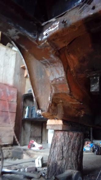 Fragment podwozia samochodu w trakcie naprawy, podpartego drewnianym klockiem na pniu, widoczne ślady korozji i napraw blacharskich.