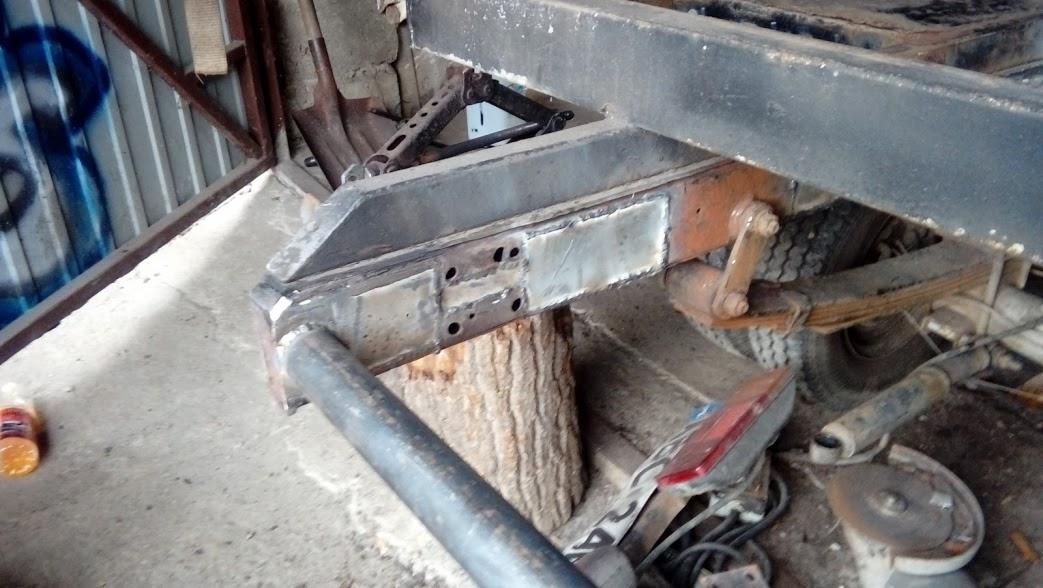 Fragment ramy pojazdu po naprawach blacharskich, z widocznymi łatami i spawami, umieszczony na pniu drzewa, obok leżą szlifierka kątowa i lampa.