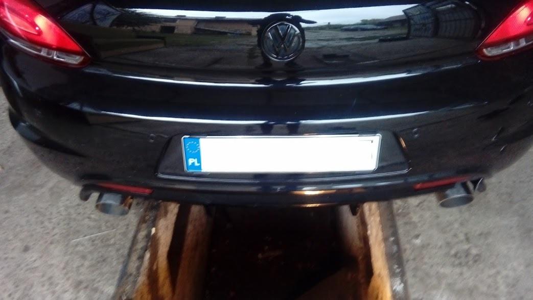 Czarny samochód Volkswagen Scirocco z polską tablicą rejestracyjną, widok od tyłu, z widocznymi dwoma rurami wydechowymi, nad kanałem samochodowym.