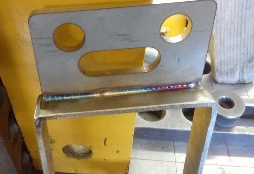 Metalowy element konstrukcyjny z widocznym spoiwem spawalniczym o opalizujących kolorach, z trzema otworami (dwa okrągłe i jeden owalny), spawany do profilu kwadratowego, na tle żółtej konstrukcji.