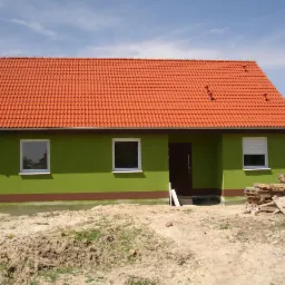 Niskoenergetyczny, parterowy dom murowany z czerwoną dachówką i zieloną elewacją w trakcie wykończenia, widoczne palety i elementy budowlane przed wejściem.