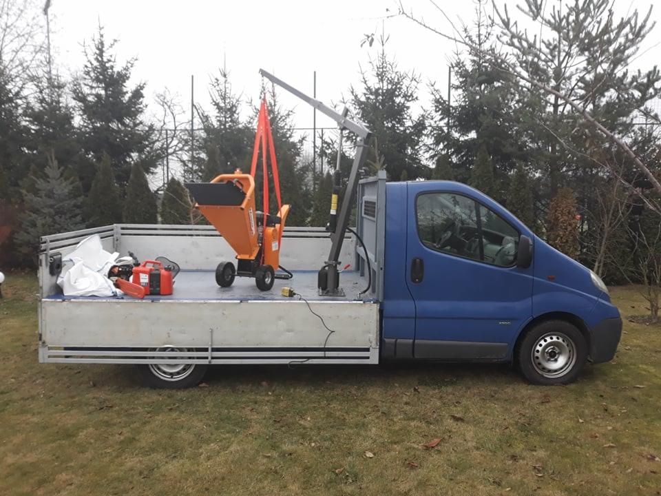 Niebieski samochód dostawczy z podnośnikiem transportuje pomarańczowy rozdrabniacz do gałęzi, na platformie leżą worki i piła spalinowa, w tle widoczne drzewa.