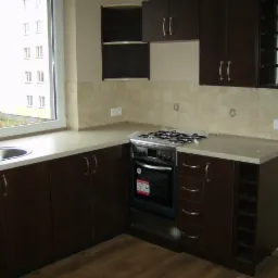 Nowoczesna, brązowa zabudowa kuchenna z blatem w jasnym odcieniu w mieszkaniu w Katowicach, widok na okno i budynek na zewnątrz.