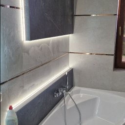 Mateusz Kaleta - Wykończona łazienka z narożną wanną, szarymi płytkami imitującymi kamień, podświetleniem LED i drewnianą szafką pod umywalką. Widoczne detale wykończeniowe.