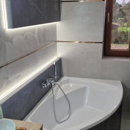 Mateusz Kaleta - Wykończona łazienka z narożną wanną obudowaną ciemnymi płytkami imitującymi kamień. Jasne płytki na ścianach z dekoracyjnymi, złotymi listwami. Okno z drewnianą ramą.