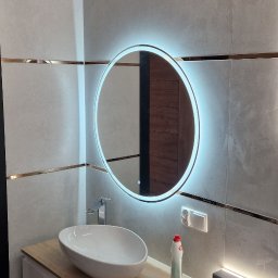 Mateusz Kaleta - Nowoczesna łazienka z owalną umywalką nablatową, szafką z drewnianym blatem i okrągłym lustrem z podświetleniem LED. Płytki ścienne w odcieniach szarości z dekoracyjnymi listwami.