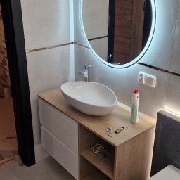 Mateusz Kaleta - Nowoczesna łazienka z owalną umywalką nablatową, podświetlanym lustrem i szafką z drewnianym blatem. Widoczne detale wykończenia i akcesoria łazienkowe.