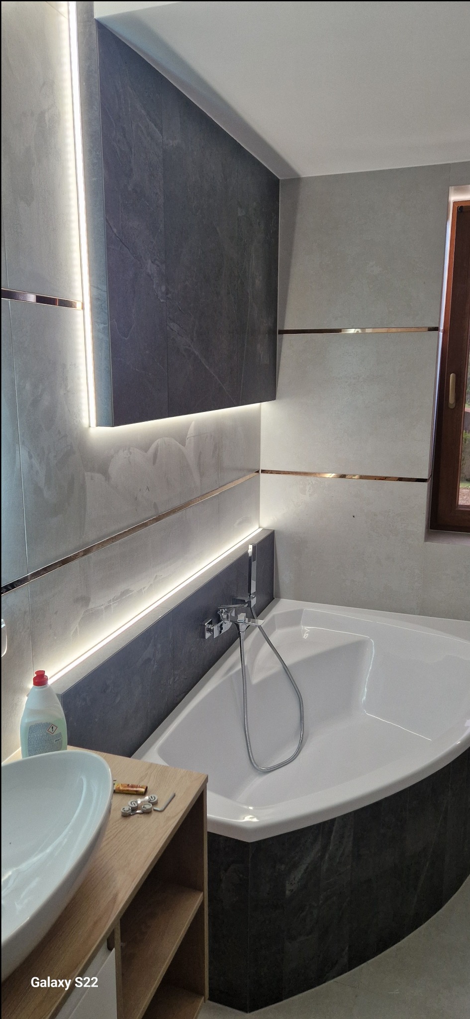 Wykończona łazienka z narożną wanną, szarymi płytkami imitującymi kamień, podświetleniem LED i drewnianą szafką pod umywalką. Widoczne detale wykończeniowe.