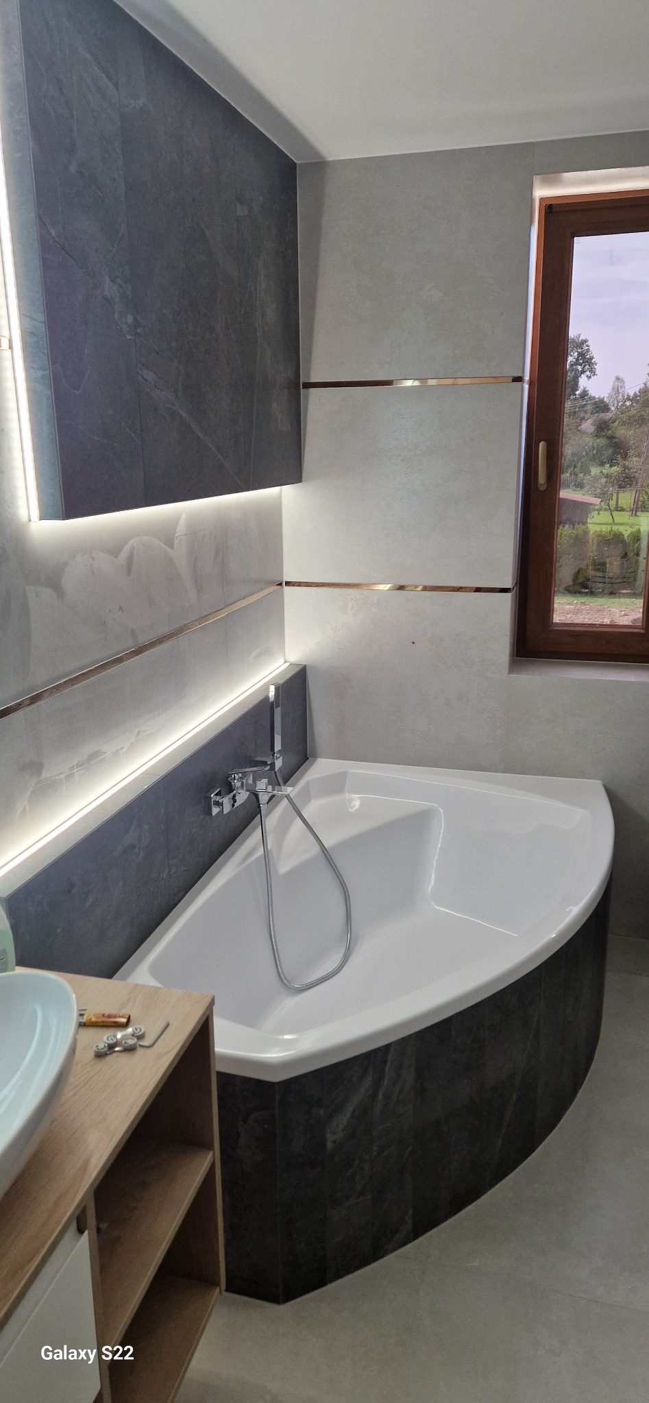 Wykończona łazienka z narożną wanną obudowaną ciemnymi płytkami imitującymi kamień. Jasne płytki na ścianach z dekoracyjnymi, złotymi listwami. Okno z drewnianą ramą.