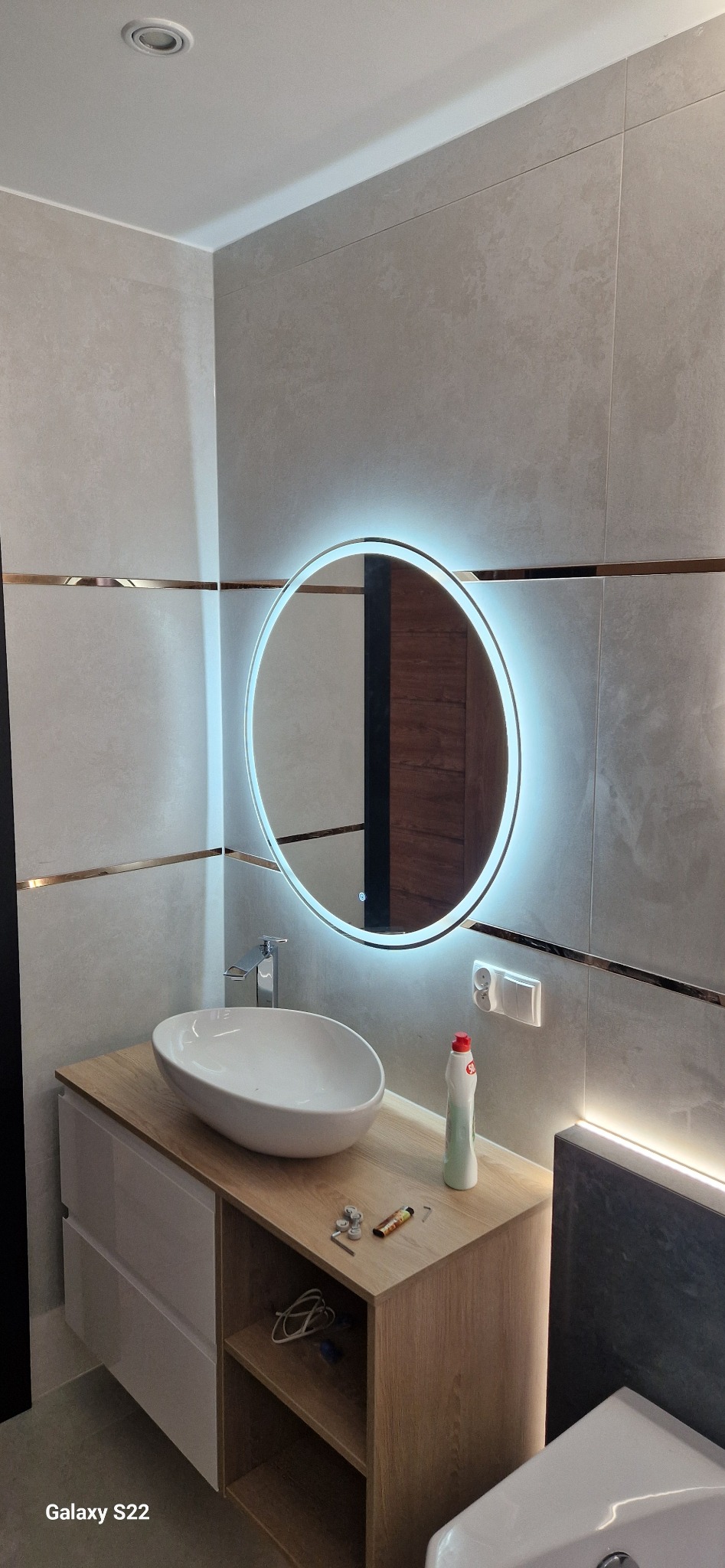 Nowoczesna łazienka z owalną umywalką nablatową, szafką z drewnianym blatem i okrągłym lustrem z podświetleniem LED. Płytki ścienne w odcieniach szarości z dekoracyjnymi listwami.
