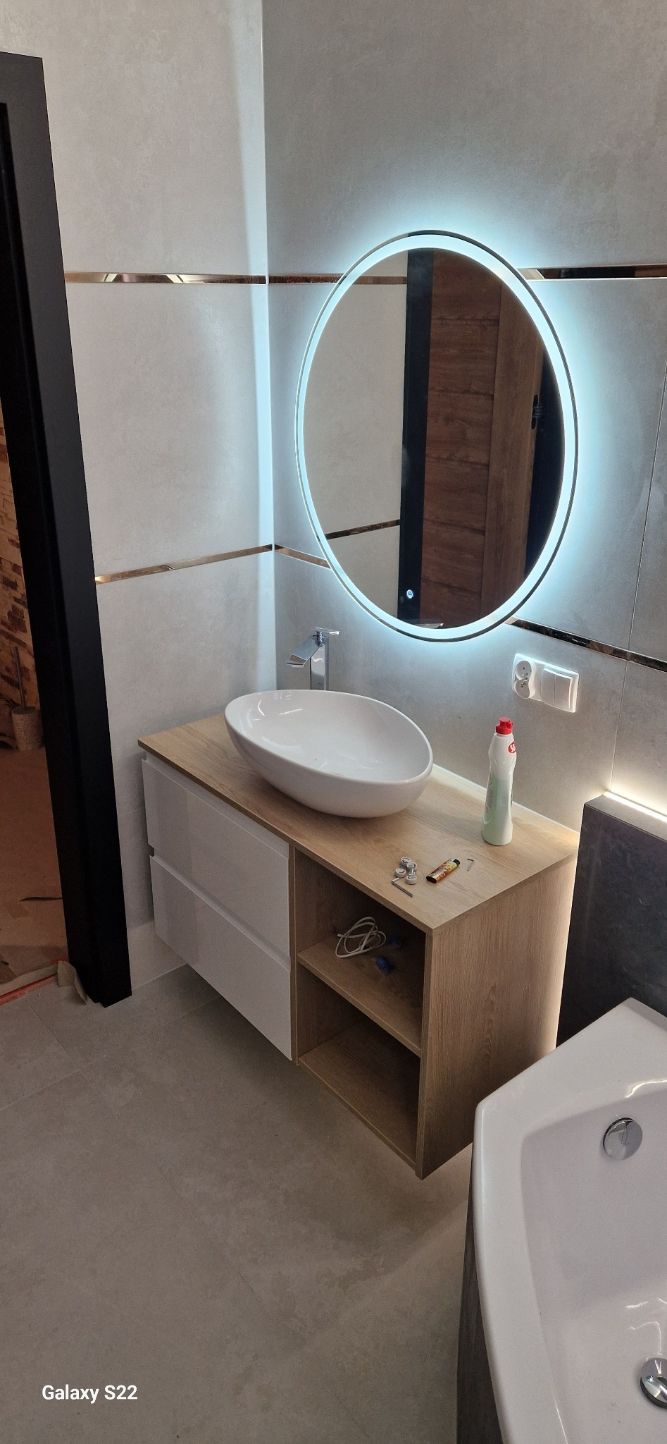 Nowoczesna łazienka z owalną umywalką nablatową, podświetlanym lustrem i szafką z drewnianym blatem. Widoczne detale wykończenia i akcesoria łazienkowe.