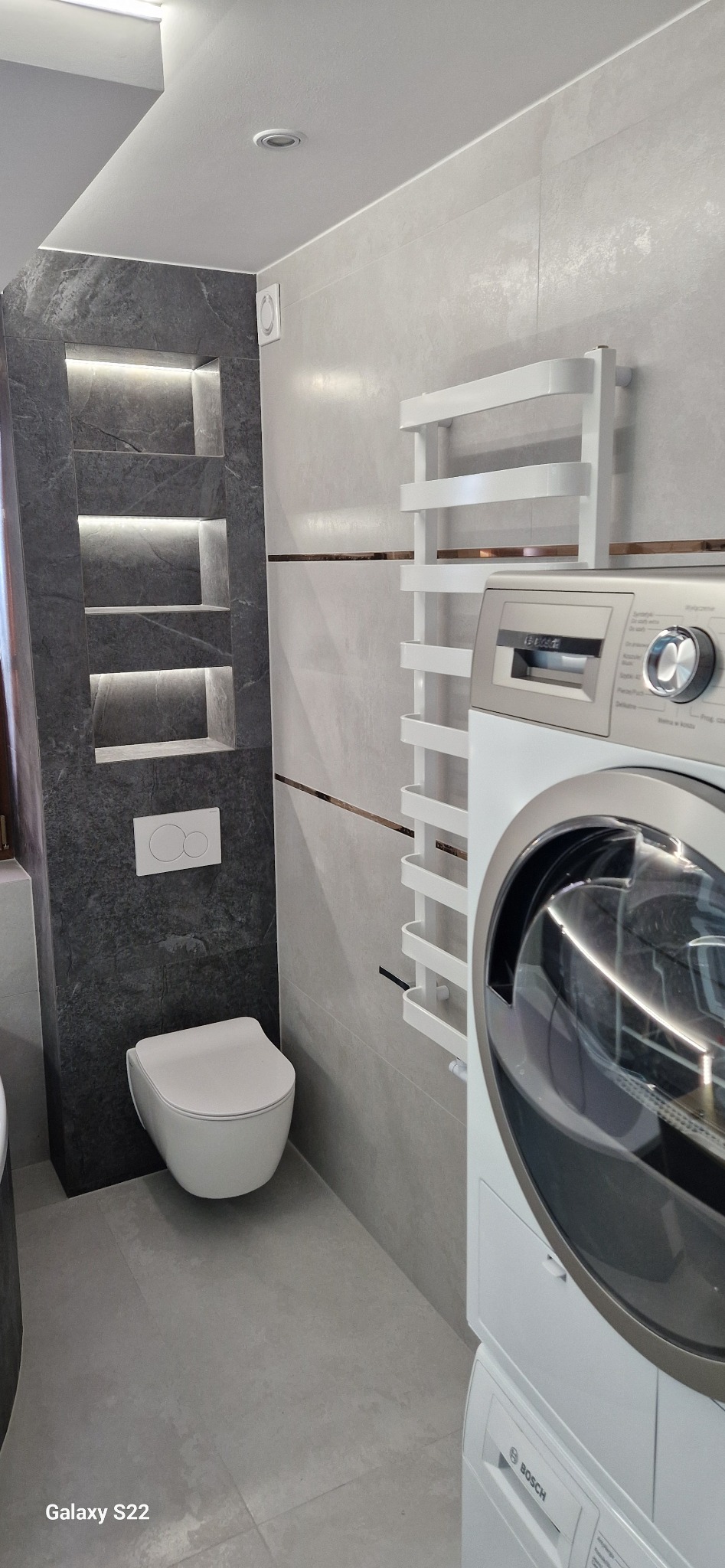 Nowoczesna łazienka w szarościach z podświetlanymi wnękami z ciemnego kamienia, wiszącą toaletą i grzejnikiem drabinkowym obok pralki.