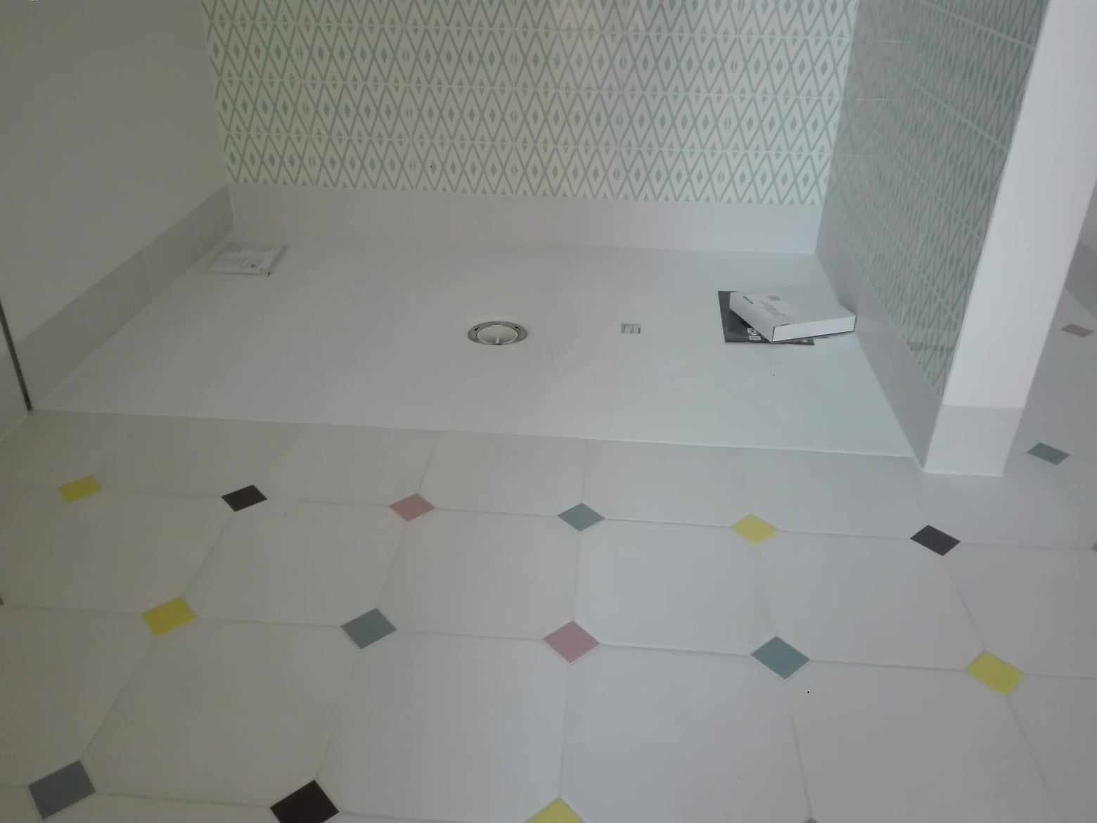 Wykończona łazienka z białymi płytkami podłogowymi z kolorowymi rombami i wzorzystymi płytkami ściennymi. Widoczny odpływ liniowy i pudełko na podłodze.