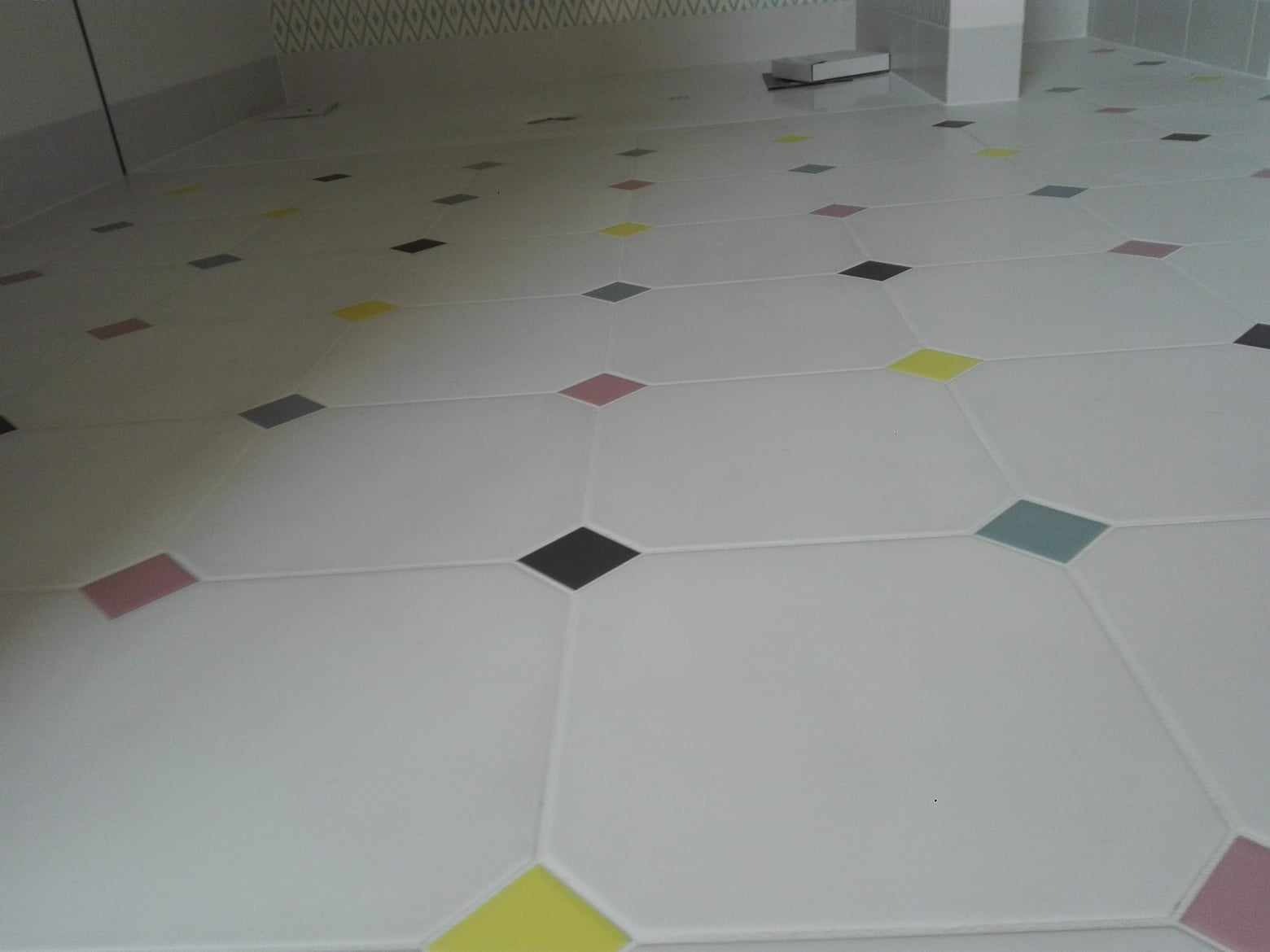 Biała podłoga z wzorem rombów w kolorach: czarnym, żółtym, różowym i szarym. Widok z góry, płytki ułożone w regularny wzór, fuga w kolorze białym.