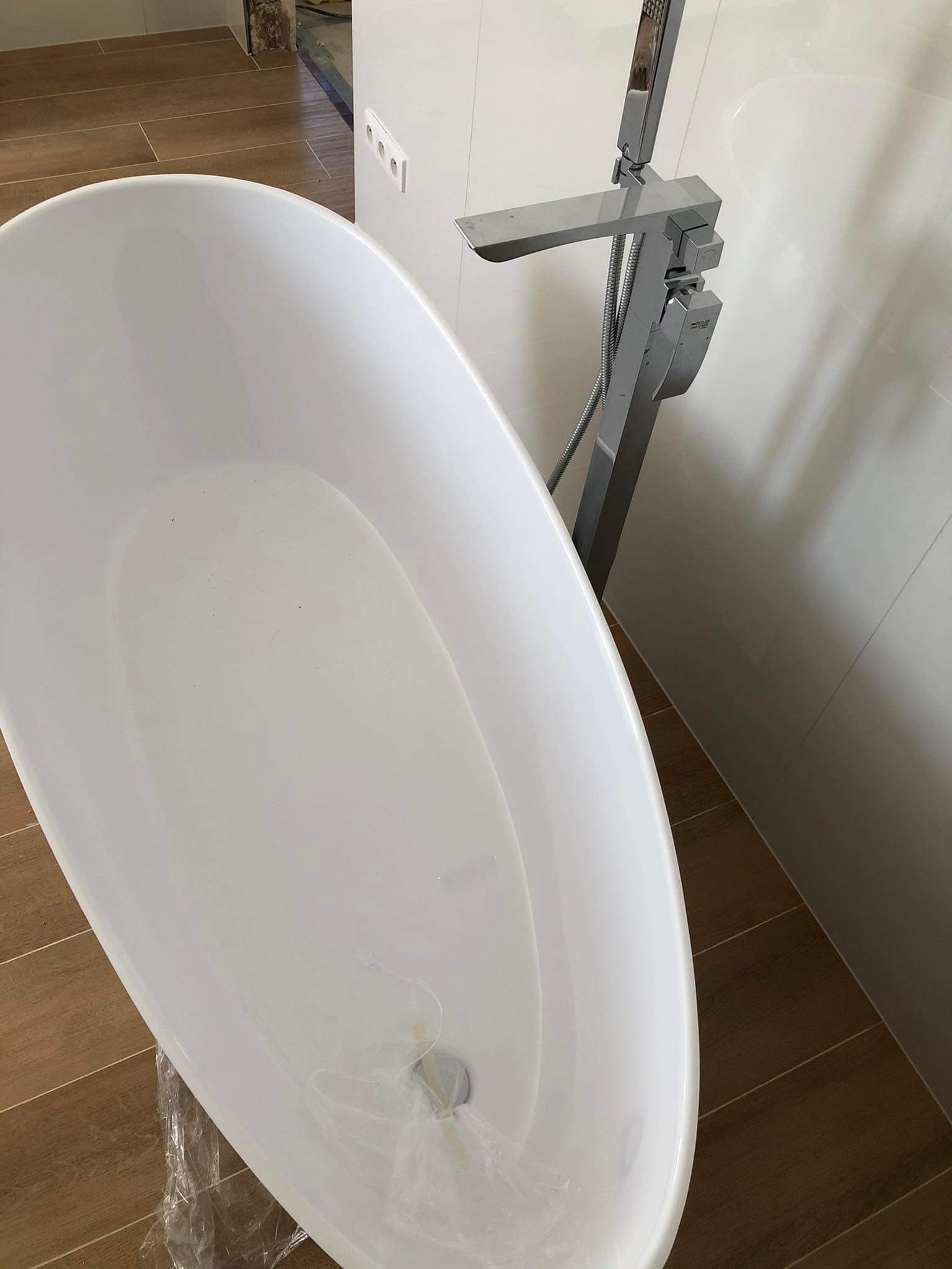 Biała, wolnostojąca wanna owalna z chromowaną baterią podłogową na tle białych płytek i brązowej podłogi. Wanna zabezpieczona folią, widoczne gniazdko elektryczne.