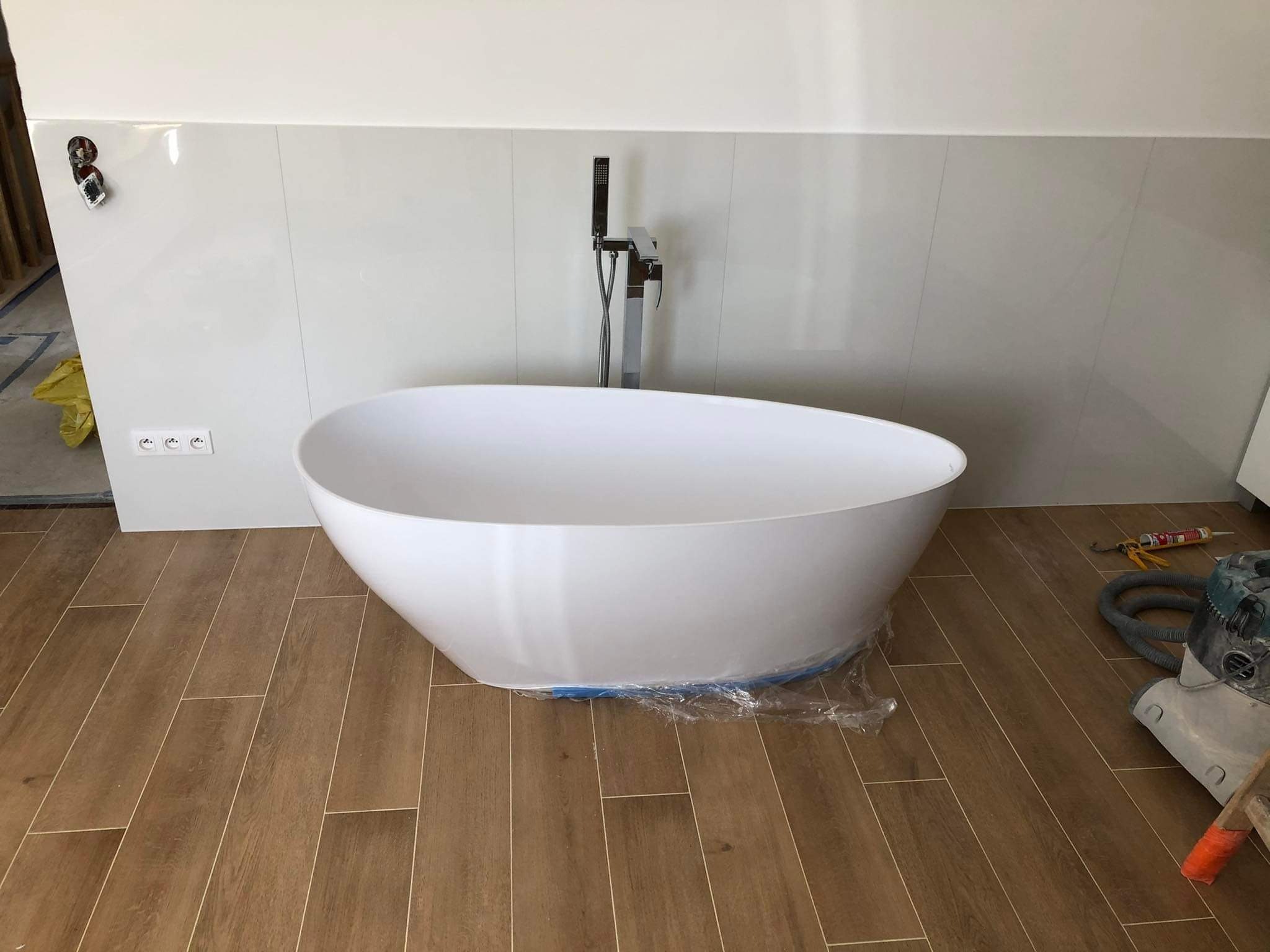 Wolnostojąca wanna owalna na drewnopodobnych płytkach w łazience w Myślenicach. Bateria podłogowa, białe panele ścienne. Widoczne narzędzia glazurnicze.