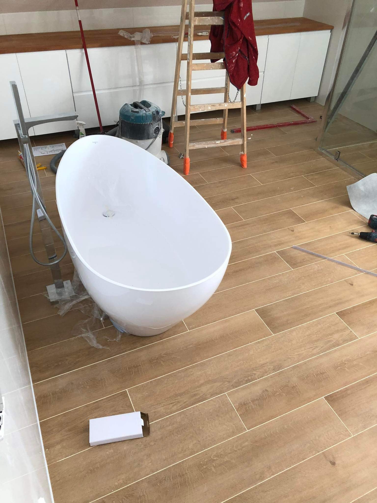 Biała wanna wolnostojąca na podłodze z płytek imitujących drewno. W tle białe szafki, drabina i narzędzia. Widoczny fragment kabiny prysznicowej.