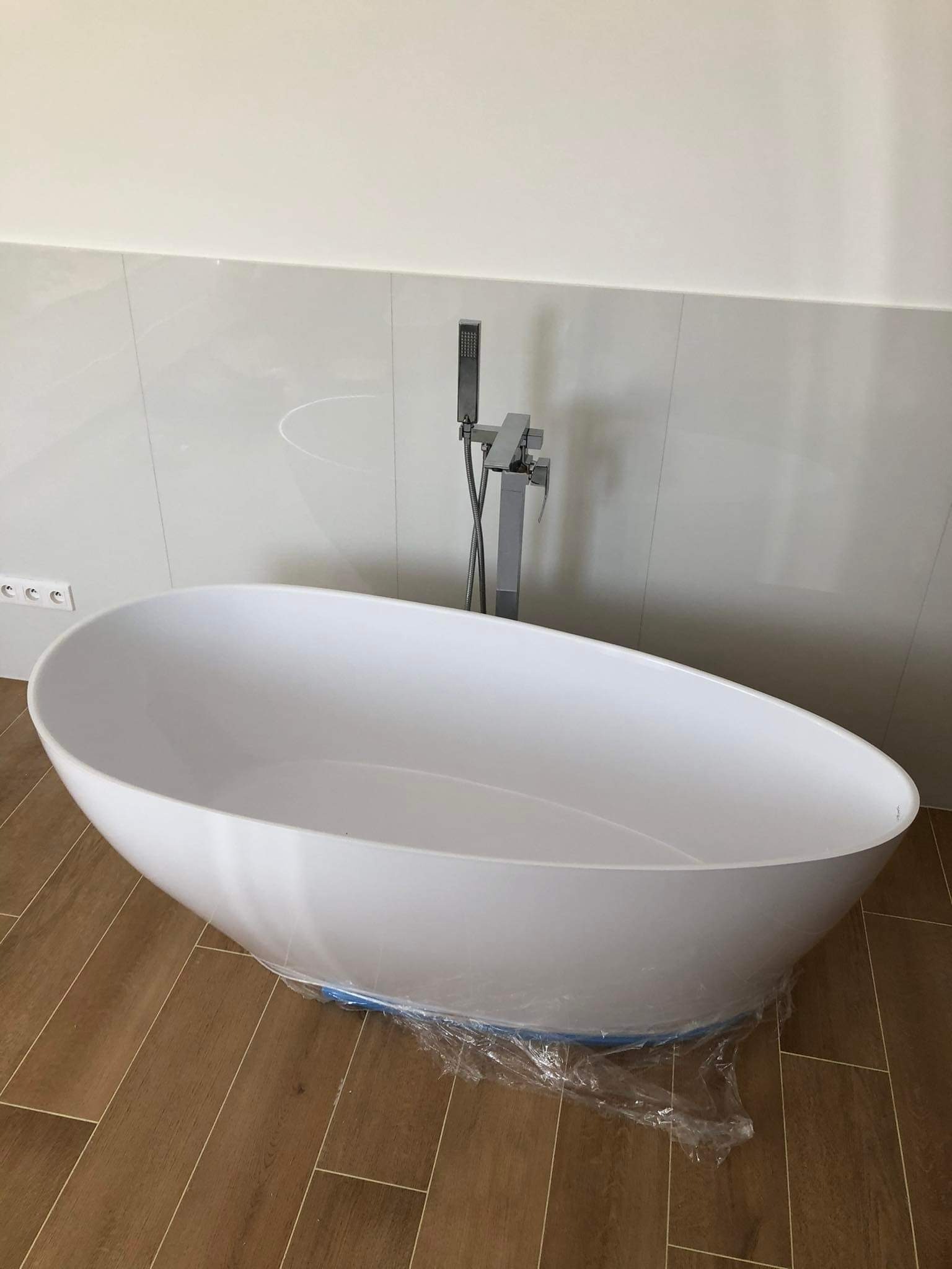 Nowoczesna, wolnostojąca wanna owalna z chromowaną armaturą na tle jasnych płytek ściennych i drewnopodobnej podłogi. Wanna zabezpieczona folią ochronną.