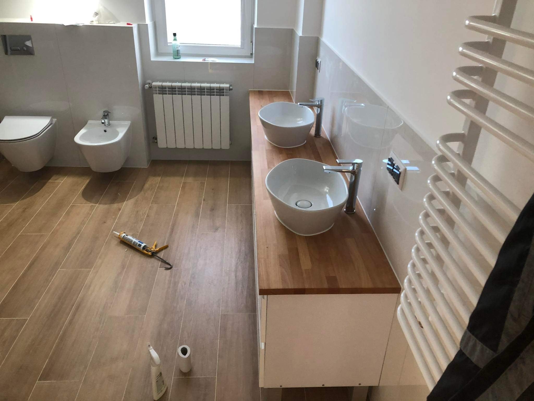 Jasna łazienka po remoncie: drewniany blat z umywalkami, beżowe płytki podłogowe i ścienne, biała armatura. Widoczne narzędzia pracy.