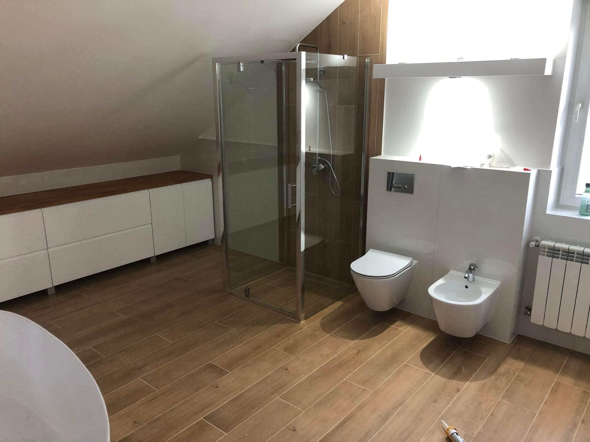 Wykończona łazienka na poddaszu ze skośnym sufitem, kabiną prysznicową, toaletą, bidetem i grzejnikiem. Podłoga wyłożona płytkami imitującymi drewno.